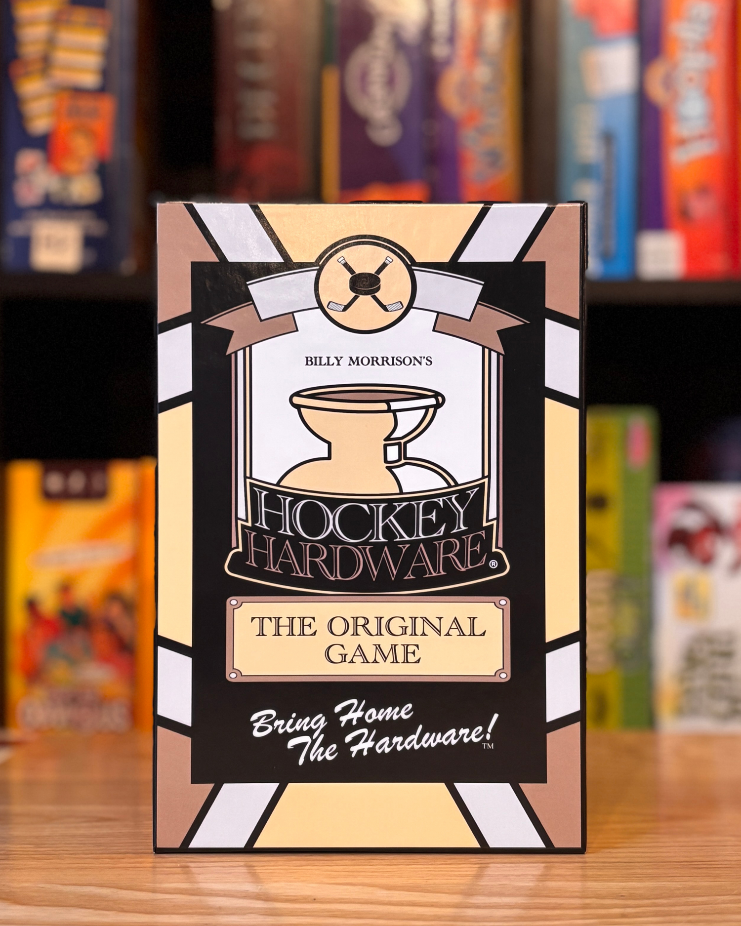 The front face of the Hockey Hardware game box positioned standing upright in front of a colourful backdrop of games on a bookshelf.